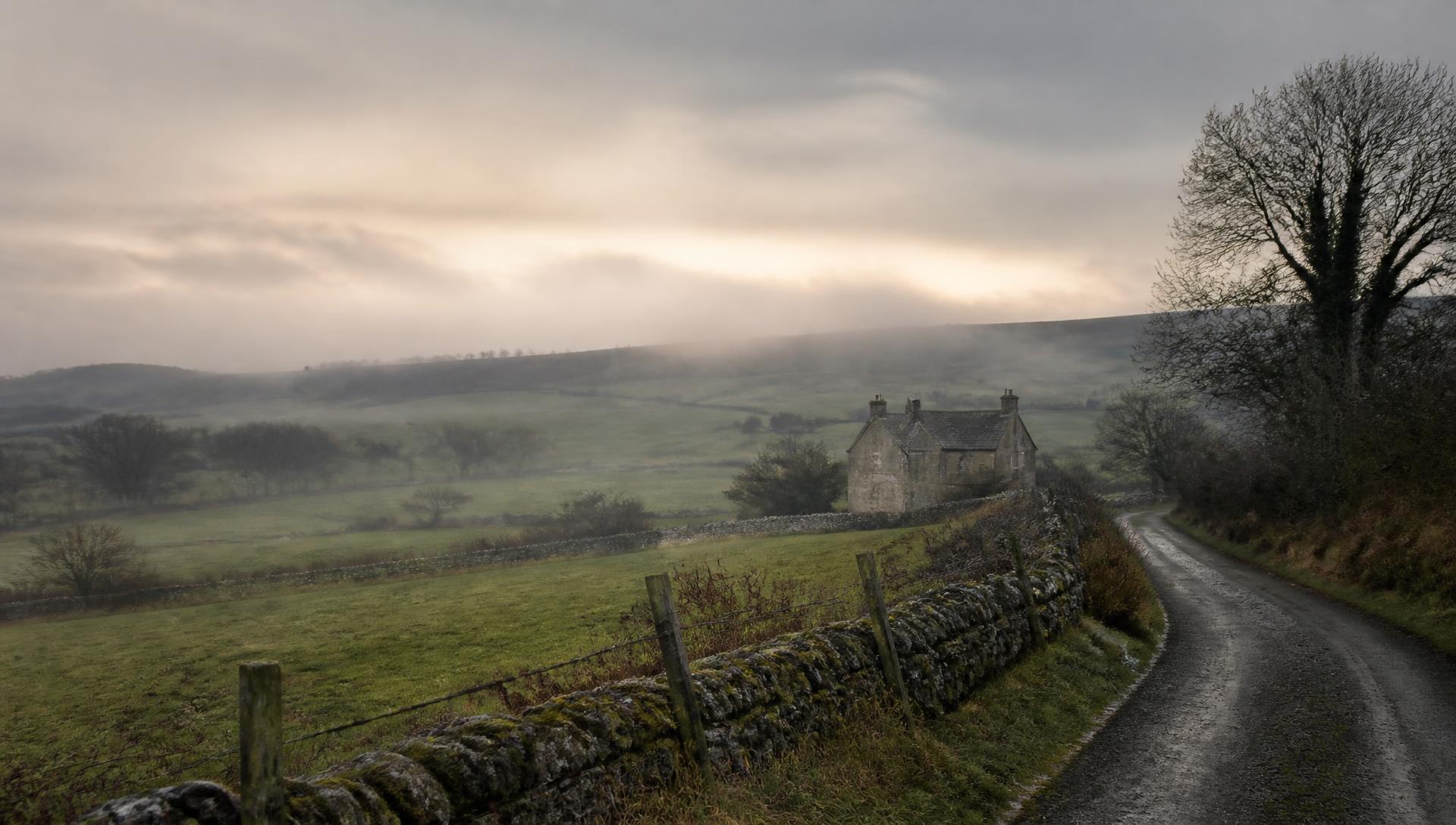 A quiet lane beside a stone wall in the Irish countryside at dawn, with a weathered farmhouse in the distance